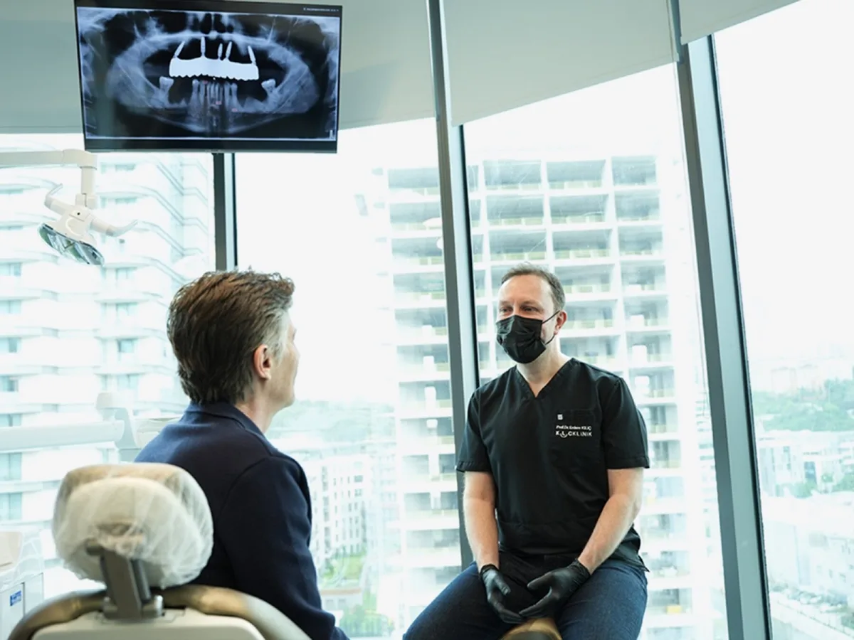 A surgeon and patient seated across from each other, with a panoramic X-ray on the wall behind them