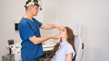 Clinician in scrubs wearing a head lamp examining a seated patient's nose during a facial aesthetic consultation
