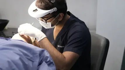 Clinician wearing a magnifying head lamp treating a draped patient's head in a clinic room