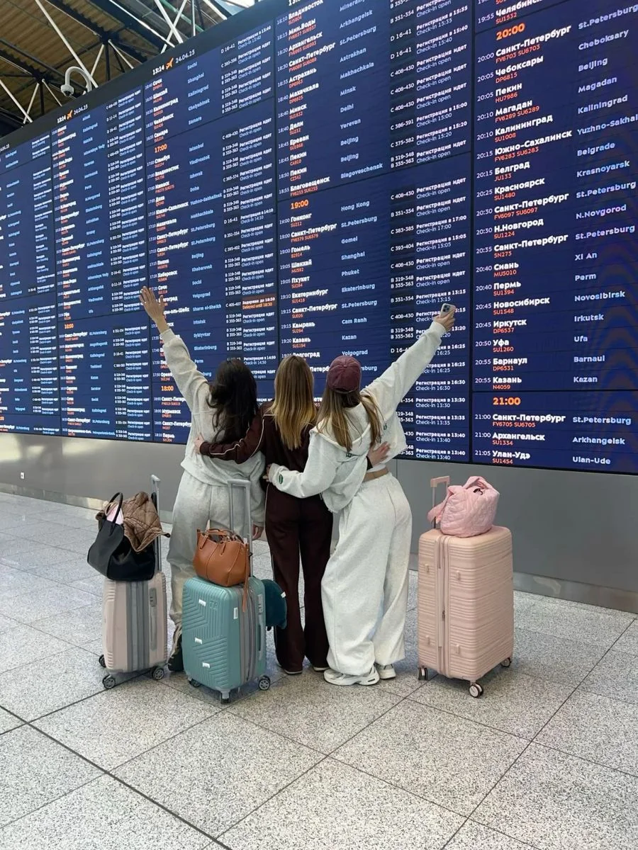 Three women with luggage waiting beneath an airport departures board